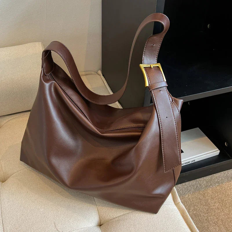 Brown leather shoulder bag with gold buckle resting on beige cushioned chair near black shelf