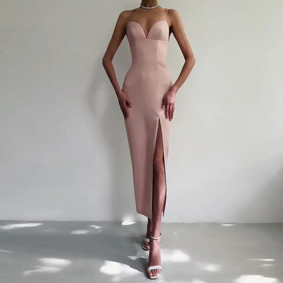 Slim woman wearing a blush pink bodycon midi dress with spaghetti straps and thigh slit, paired with white heeled sandals