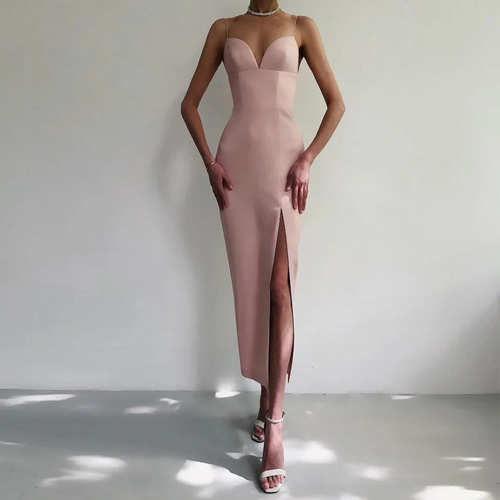 Slim woman wearing a blush pink bodycon midi dress with spaghetti straps and thigh slit, paired with white heeled sandals