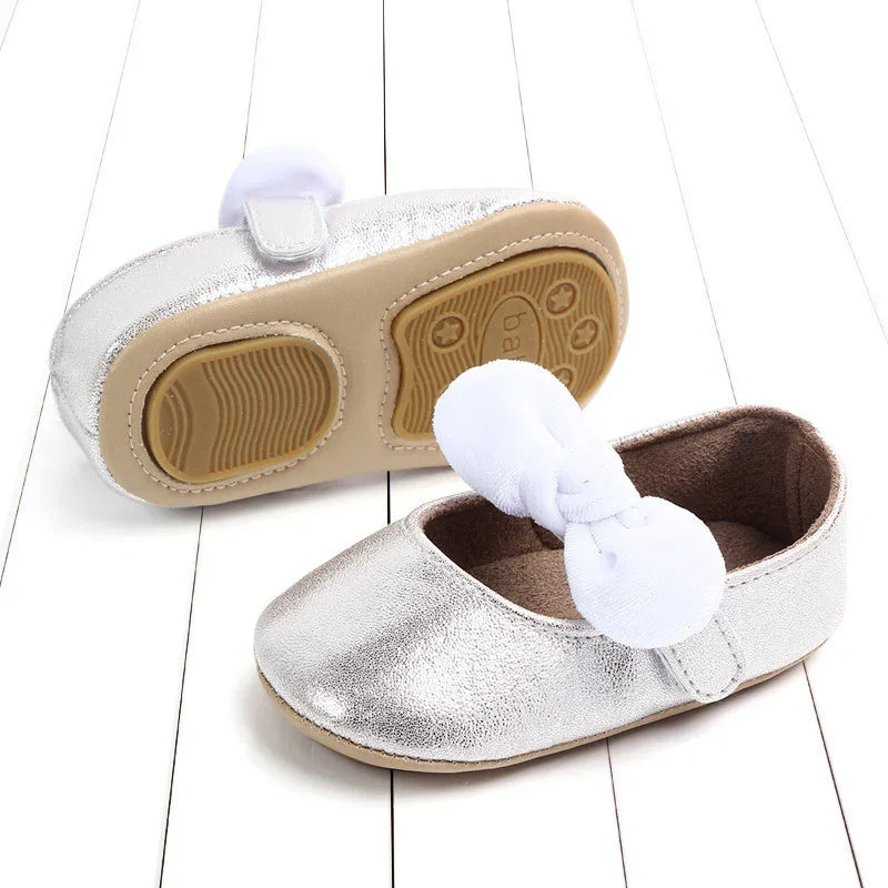 Silver baby shoes with white bow, soft sole, placed on white wooden floor