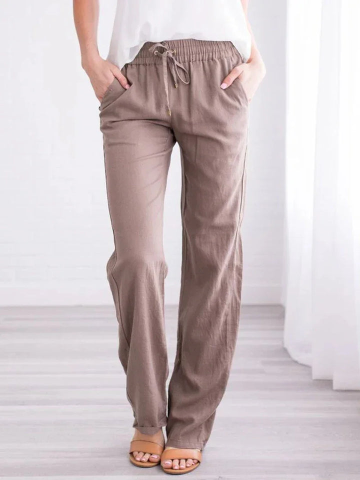 Woman wearing taupe drawstring linen pants with pockets and tan sandals in bright room