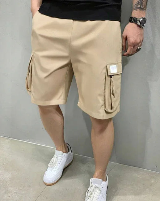 Man wearing beige cargo shorts with flap pockets, white sneakers, black t-shirt, standing indoors