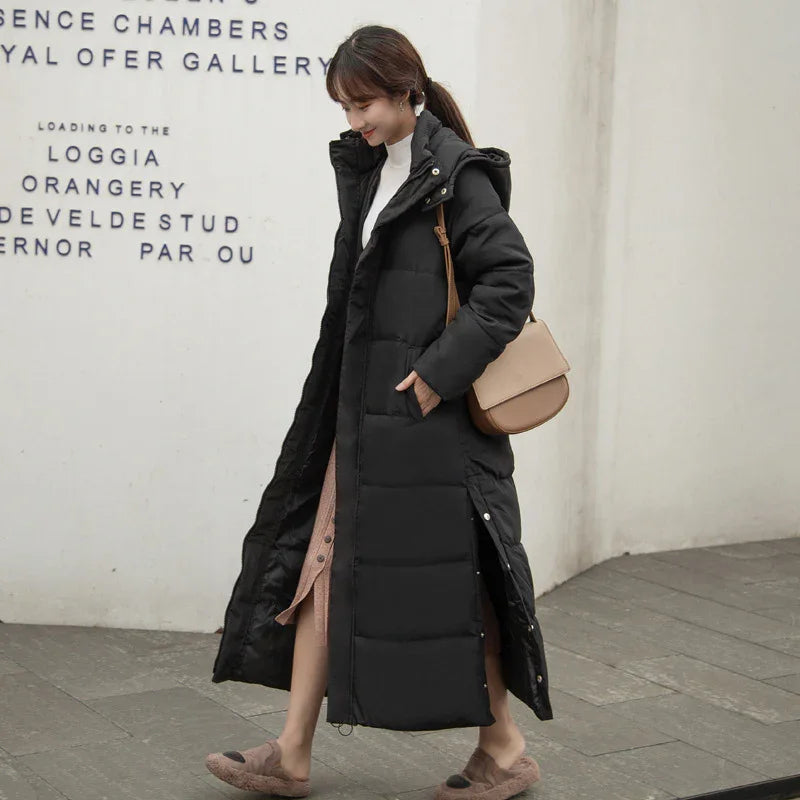 Woman wearing long black padded winter coat with hood walking outdoors carrying beige shoulder bag