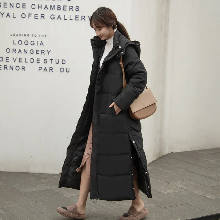 Woman wearing long black padded winter coat with hood walking outdoors carrying beige shoulder bag