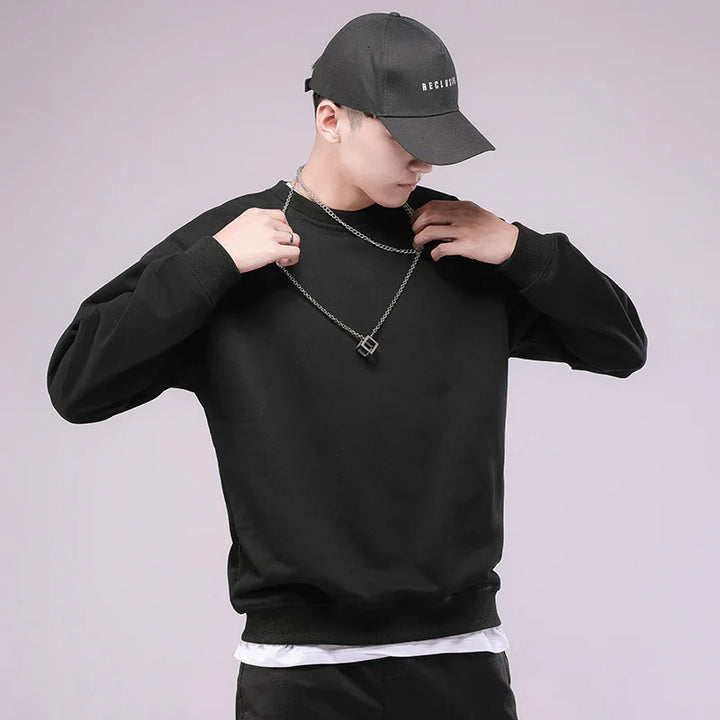 Young man wearing black sweatshirt and black cap, adjusting silver pendant necklace on plain background