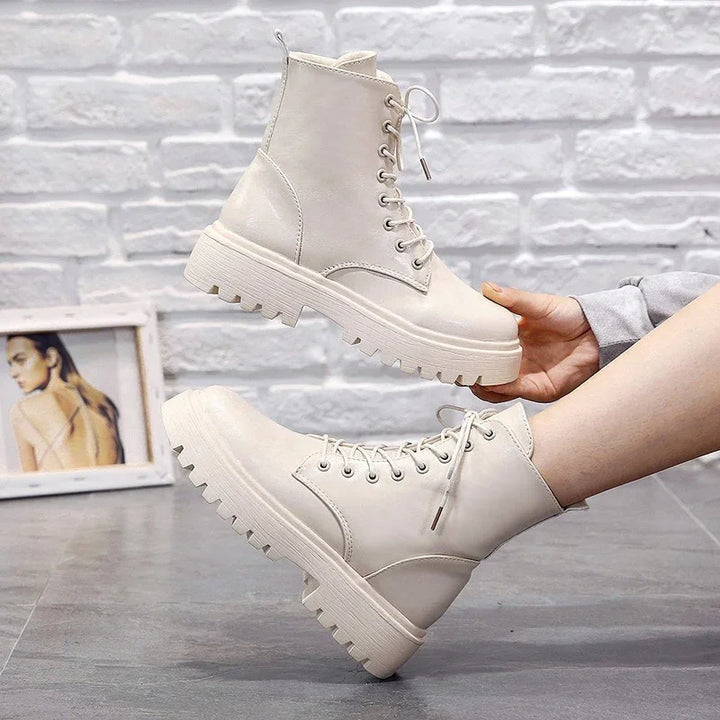 White lace-up combat boots with chunky soles worn on feet against a gray floor and white brick wall background