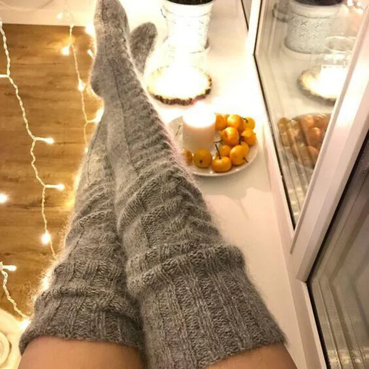 Cozy gray knit knee-high socks on legs next to warm string lights and a plate with orange fruits and candle