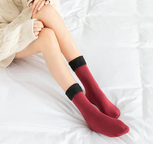 Woman wearing cozy maroon socks with black cuffs, sitting on white bed sheets in beige sweater