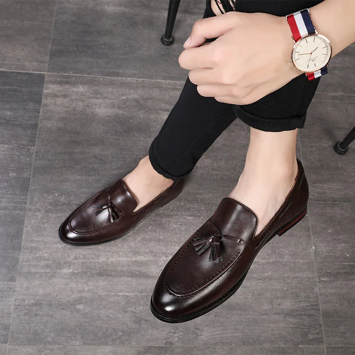 Male wearing dark brown leather tassel loafers with black pants and a wristwatch on tiled floor