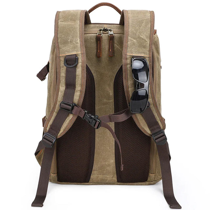 Brown canvas backpack with padded mesh back, adjustable straps, and black sunglasses clipped on shoulder strap