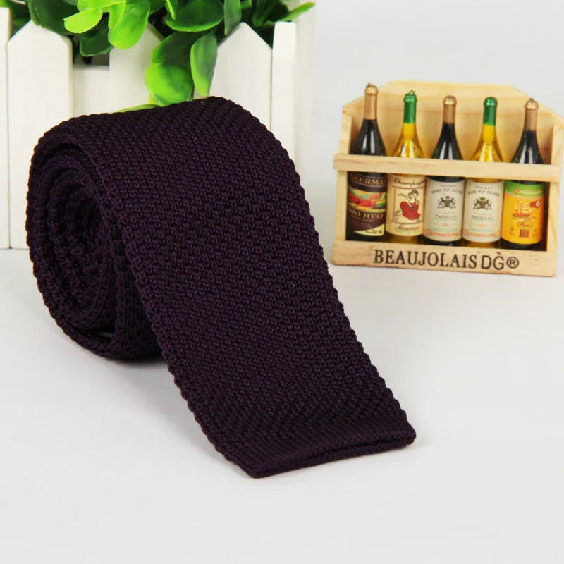 Dark purple knitted tie rolled up on white surface with miniature wooden wine bottle crate and green plants background
