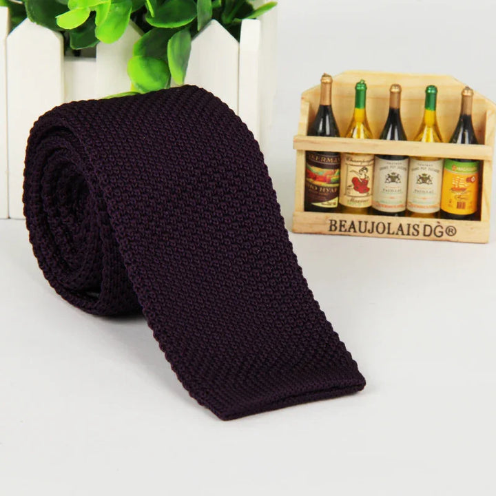 Dark purple knitted tie rolled up on white surface with miniature wooden wine bottle crate and green plants background