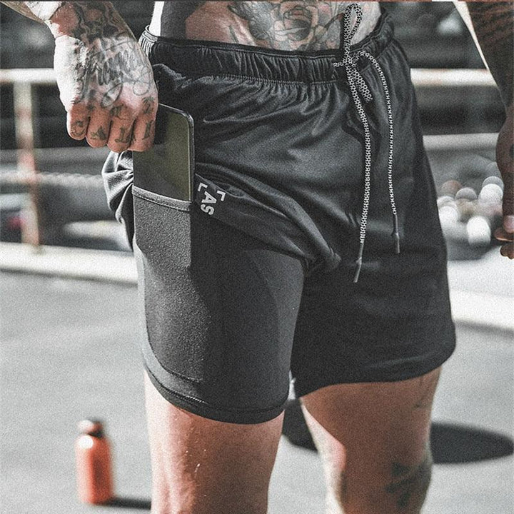 Close-up of tattooed man wearing black athletic shorts with side pocket holding a smartphone outdoors