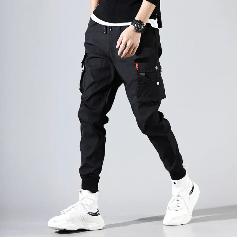 Man wearing black cargo jogger pants with white sneakers and black wristwatch on gray background