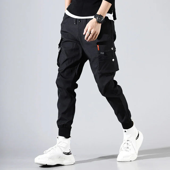 Man wearing black cargo jogger pants with white sneakers and black wristwatch on gray background