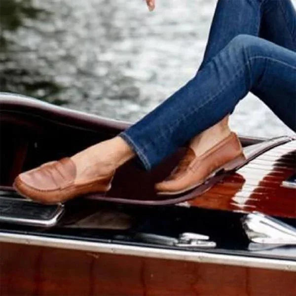Person wearing brown leather loafers and blue jeans sitting on a wooden boat near water