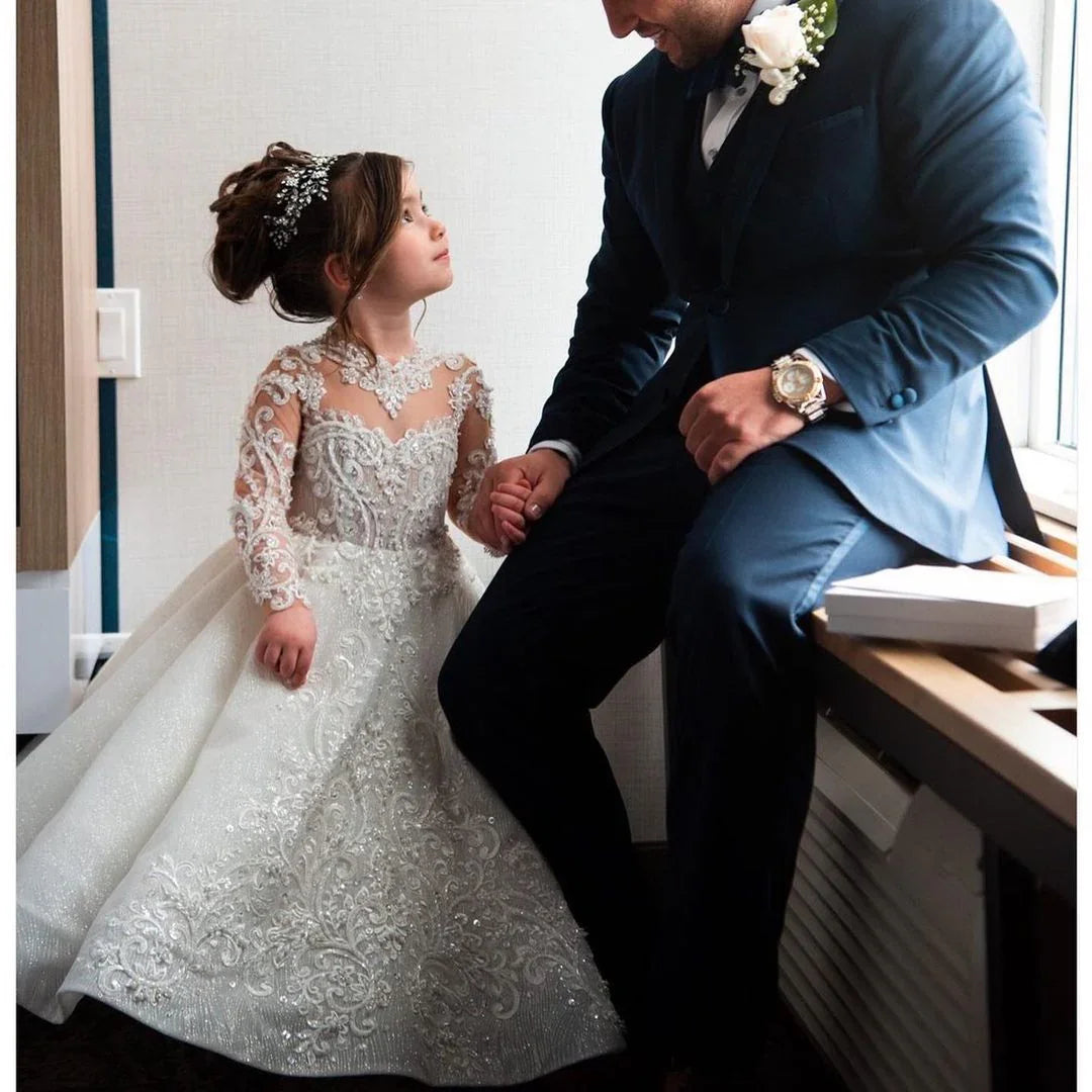 Little girl in embellished white lace dress holding hands with man in blue suit near window