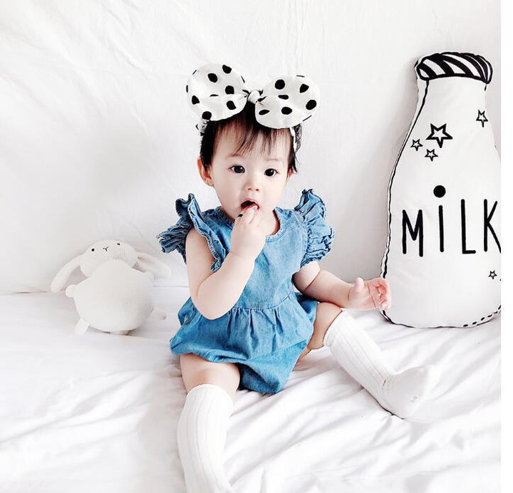 Cute baby girl in blue dress and polka dot bow headband sitting on white bed with milk bottle pillow and plush toy