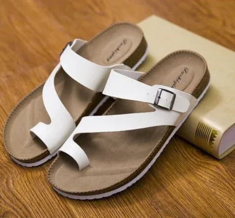 Pair of white leather buckle sandals with toe loop on tan footbed placed on wooden floor next to a closed book