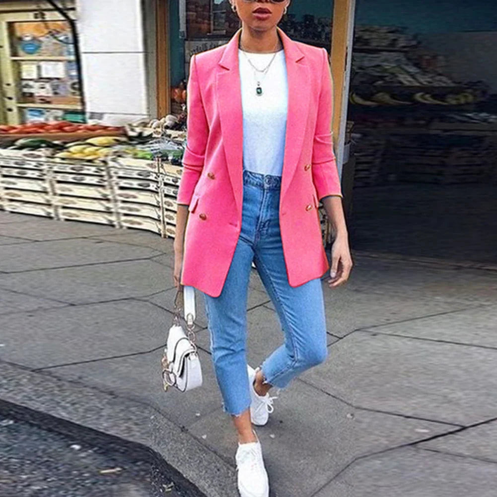 Woman wearing pink blazer, white top, blue jeans, and white sneakers standing on city sidewalk