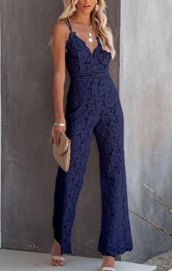 Woman wearing navy blue lace jumpsuit with spaghetti straps, holding beige clutch, standing against gray wall
