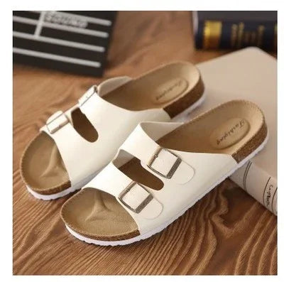 White double buckle slide sandals with cork footbed on wooden surface, casual summer footwear