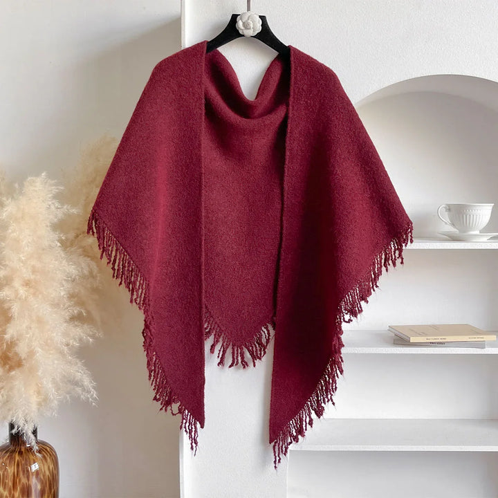 Burgundy wool poncho with fringe edges hanging on black hanger in minimalist interior