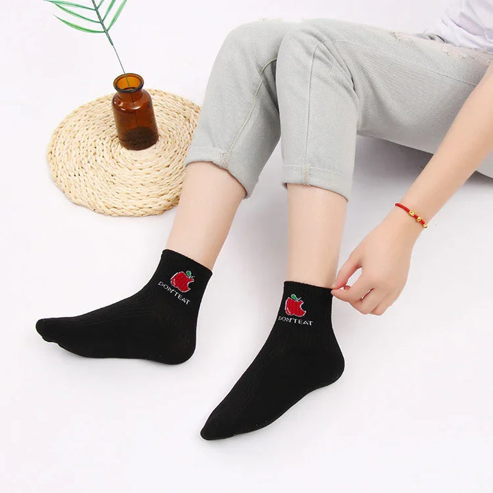 Person wearing black ankle socks with red apple and text 'DON'T EAT' sitting on white floor with brown vase on woven mat