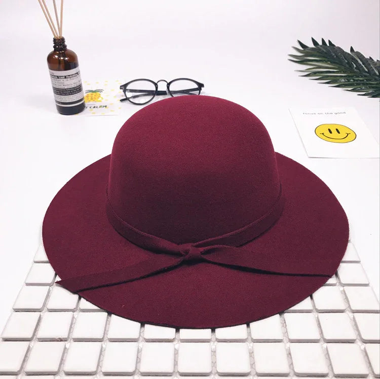 Maroon wide-brim wool felt hat with a bow band on white tiled surface, glasses, diffuser, and smiley card