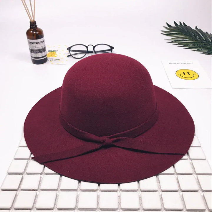 Maroon wide-brim wool felt hat with a bow band on white tiled surface, glasses, diffuser, and smiley card