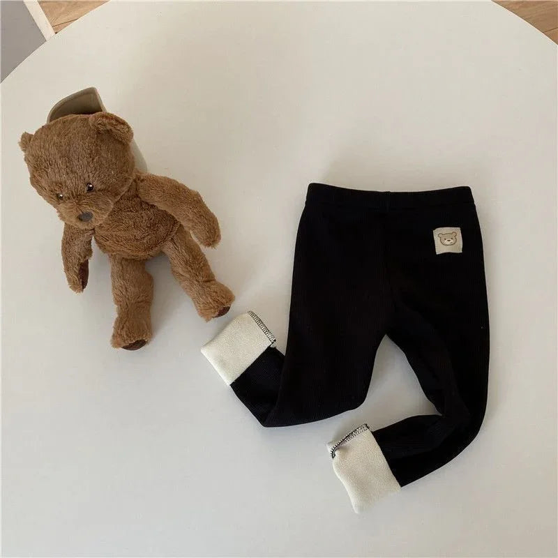 Brown teddy bear plush toy next to black baby leggings with white cuffs and teddy patch on white table