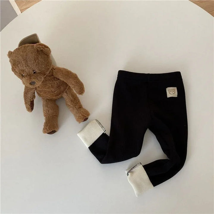 Brown teddy bear plush toy next to black baby leggings with white cuffs and teddy patch on white table