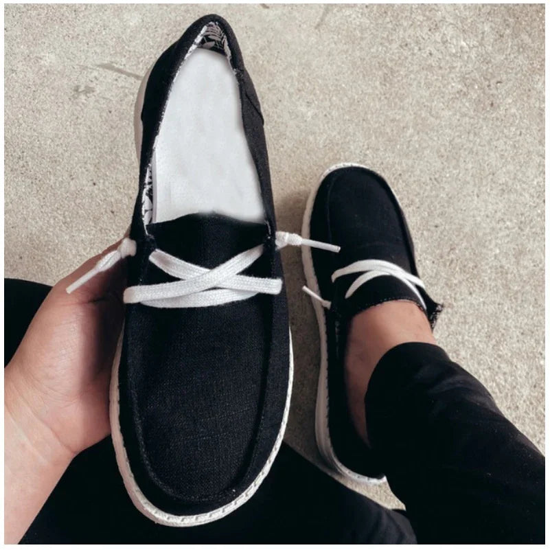 Black casual slip-on shoes with white laces and soles, one shoe held in hand and worn on foot on concrete floor