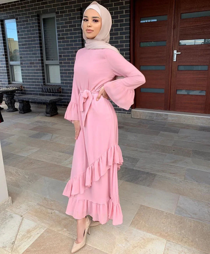 Woman wearing a pink ruffled modest dress and beige hijab standing on stone floor in front of wooden door