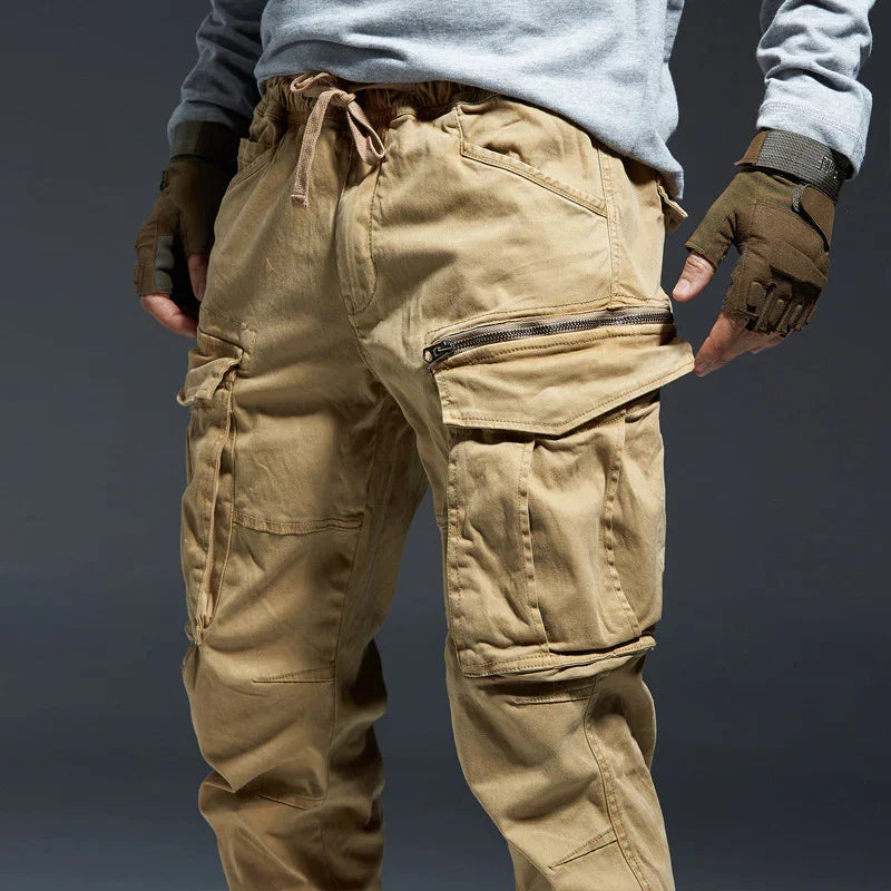 Men's beige cargo pants with multiple pockets and zipper details, styled with gray sweatshirt and tactical gloves