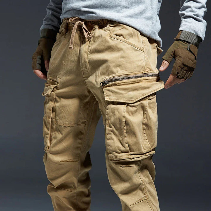 Men's beige cargo pants with multiple pockets and zipper details, styled with gray sweatshirt and tactical gloves