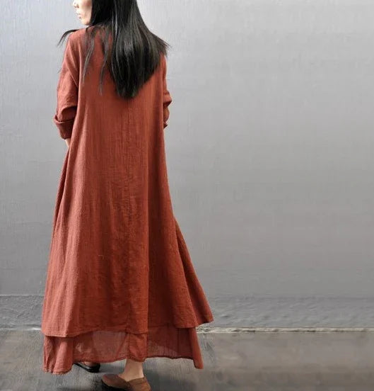 Woman wearing long rust-colored linen dress with layered hem, standing against gray wall