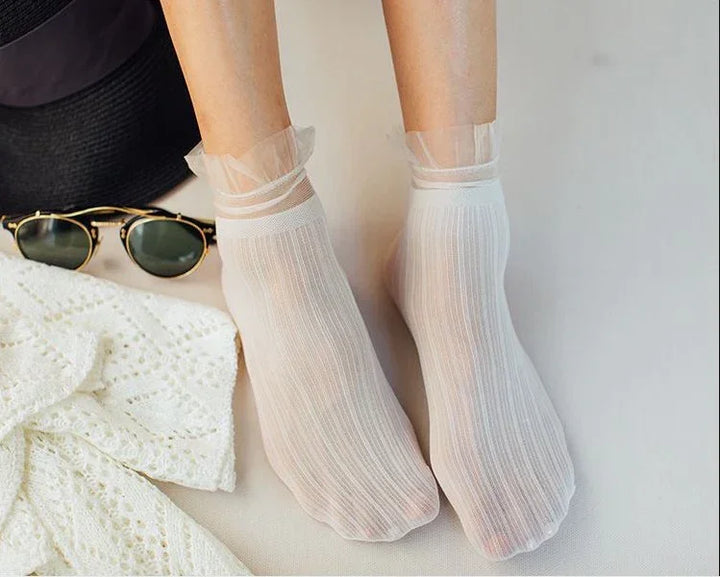 Feet wearing sheer white ankle socks with ruffled cuffs, alongside sunglasses and a white knitted garment