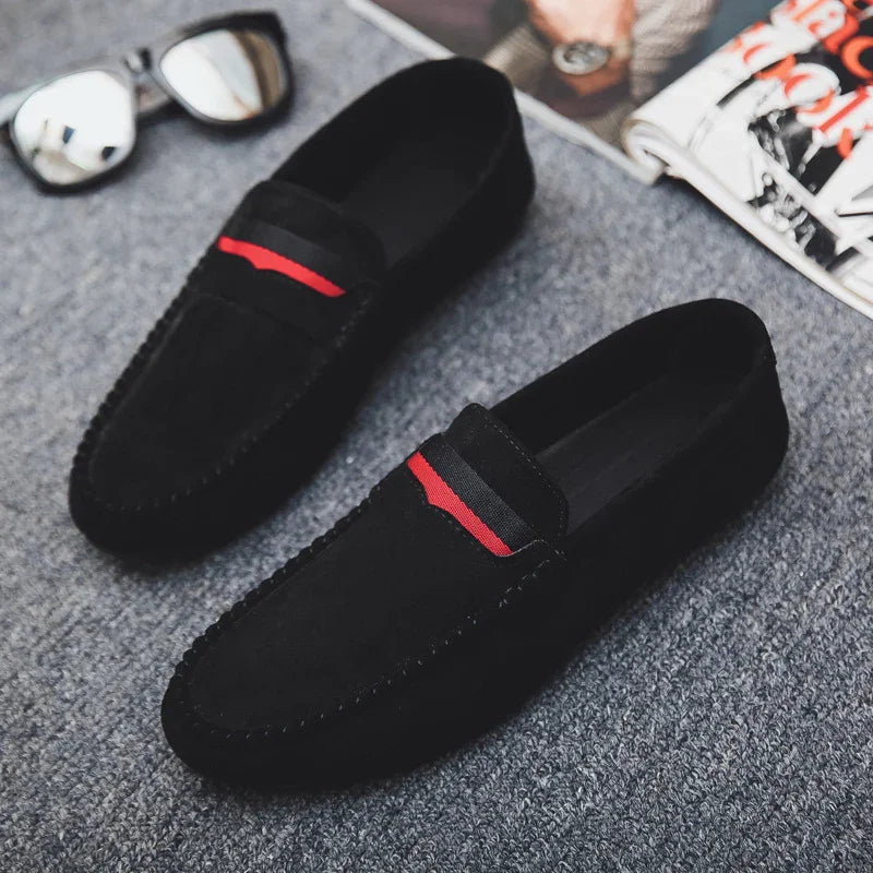Black suede men's loafers with red and black strap detail on gray carpet, sunglasses and magazine nearby