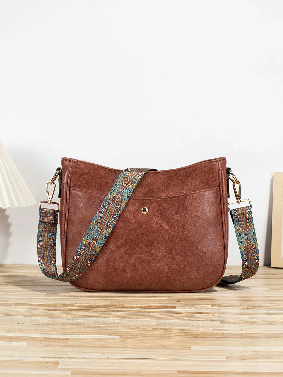Brown leather crossbody bag with colorful patterned adjustable strap on wooden surface