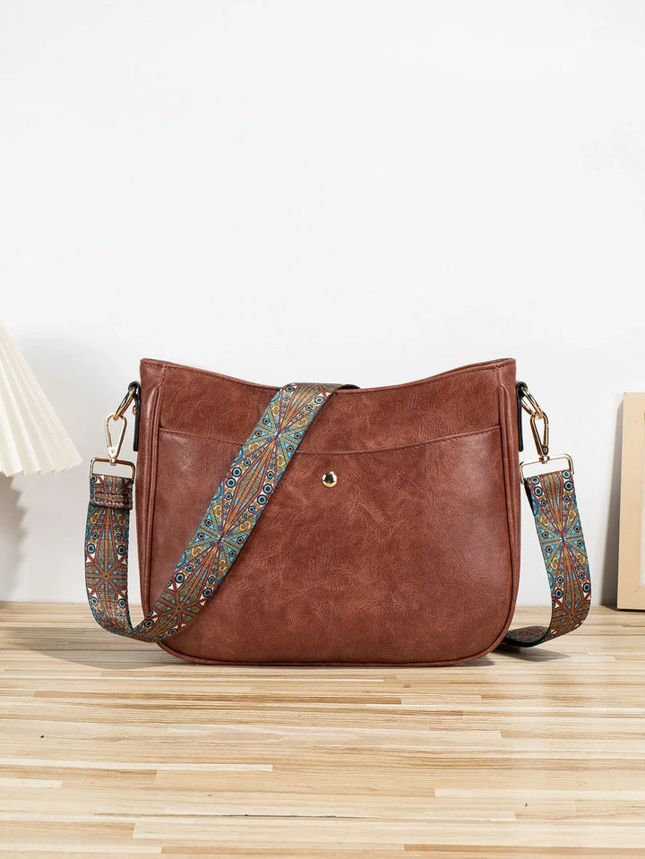 Brown leather crossbody bag with colorful patterned adjustable strap on wooden surface