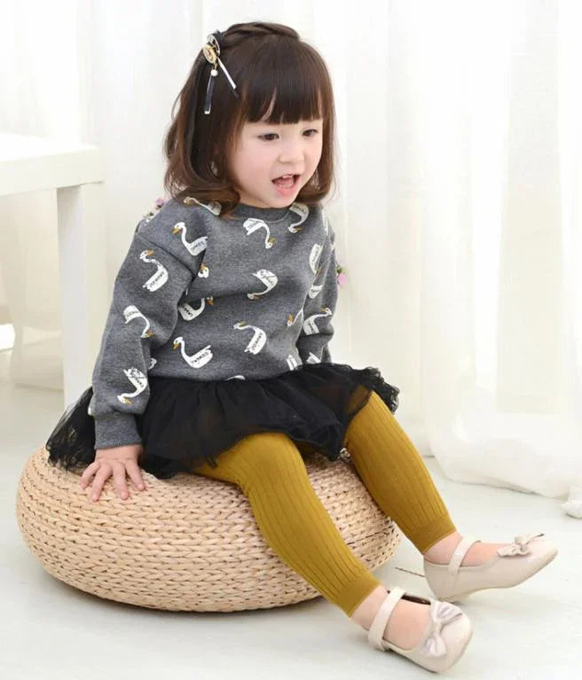 Toddler girl wearing gray swan print sweater, black tutu skirt, mustard leggings, and beige shoes sitting on a woven pouf