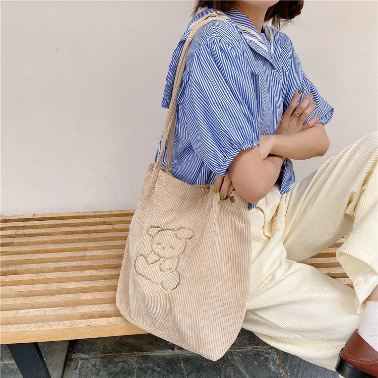Woman sitting on bench wearing blue striped shirt and cream pants, carrying beige corduroy tote bag with teddy bear design