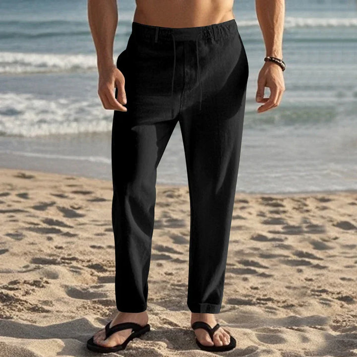 Man wearing black casual pants and black sandals standing on sandy beach near ocean