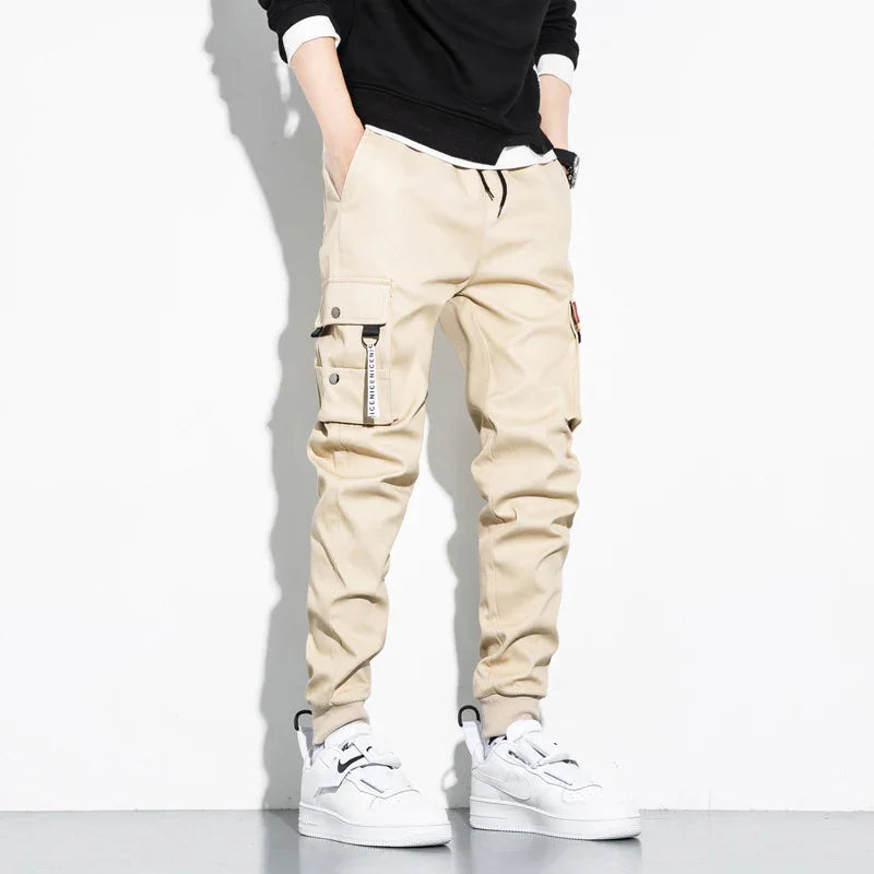 Men's beige cargo jogger pants with drawstring waist and pockets, styled with white sneakers and black sweatshirt