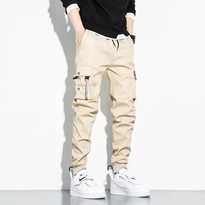 Men's beige cargo jogger pants with drawstring waist and pockets, styled with white sneakers and black sweatshirt