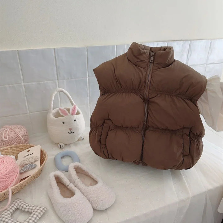Brown puffer vest on a white surface with fuzzy white baby shoes and a bunny-shaped felt bag