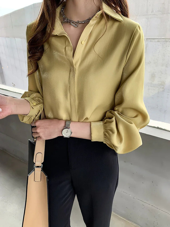 Woman wearing a shiny mustard yellow blouse with puff sleeves, black pants, silver watch, and beige handbag