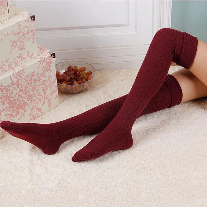 Person wearing burgundy cable knit over-the-knee socks on cream carpet near floral gift boxes and glass bowl of grapes