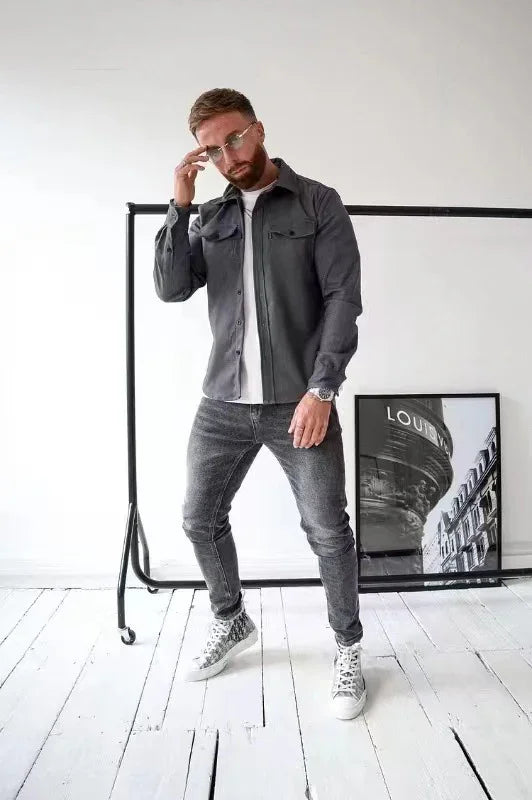 Stylish man wearing gray shirt, white t-shirt, gray jeans, and sneakers in minimalist white room with framed art